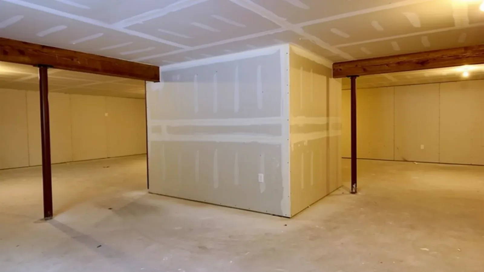 An open basement interior with a newly drywalled partition wall, its seams taped and coated, under a taped drywall ceiling