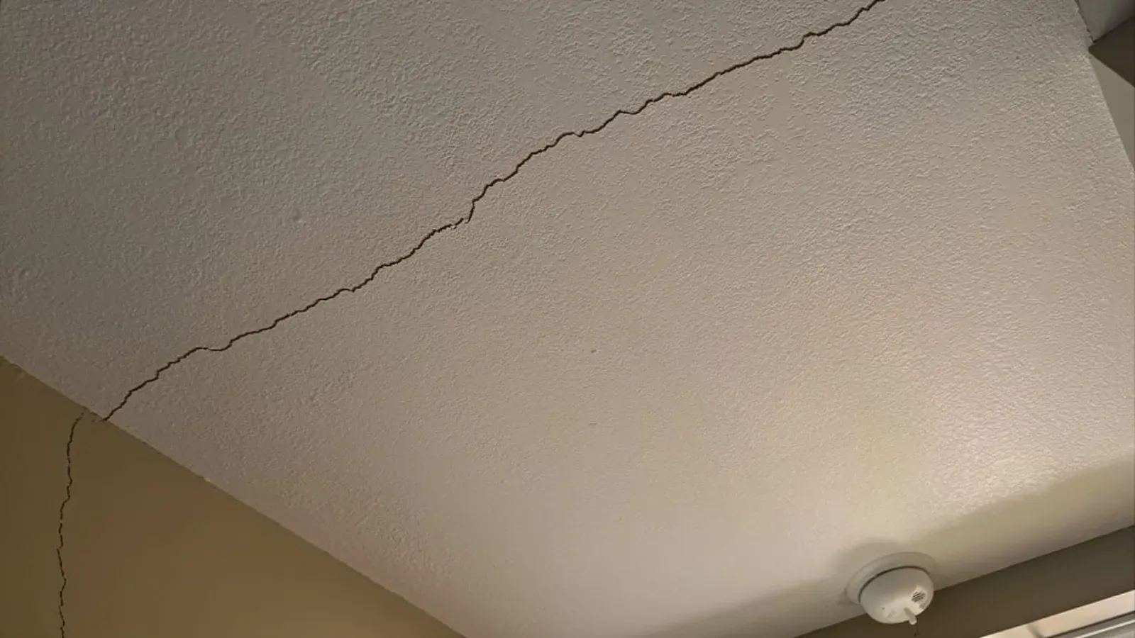 A long crack running diagonally across a lightly textured painted ceiling toward the corner where the ceiling meets the wall, with a round white smoke detector mounted nearby