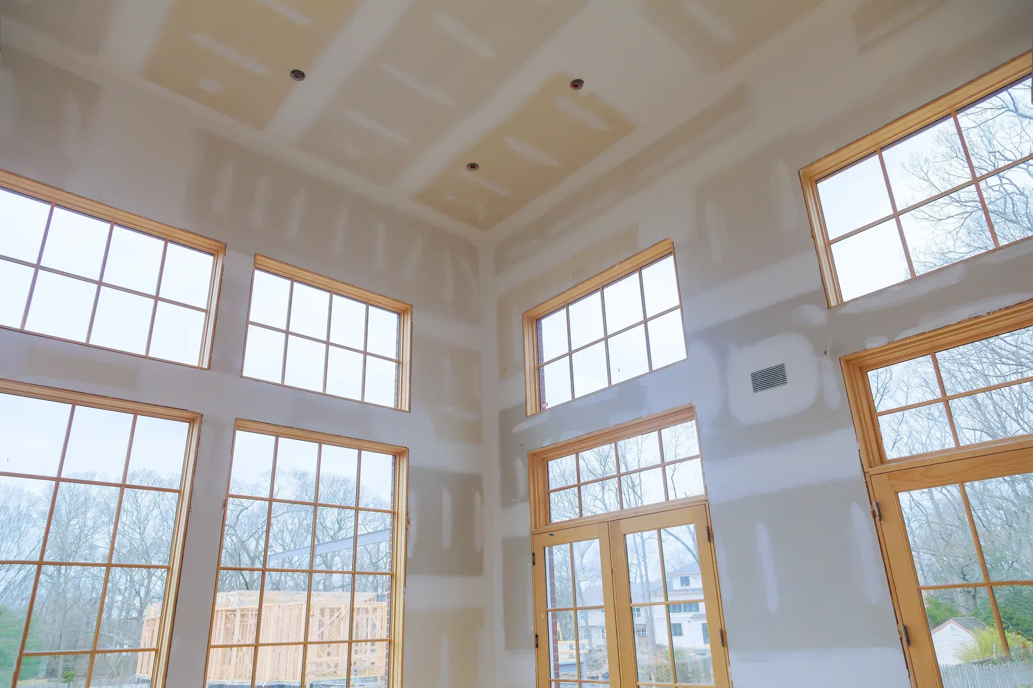 Two-story great room under construction with taped drywall walls and ceiling and a bank of tall wood-trimmed windows and French doors overlooking a wooded lot