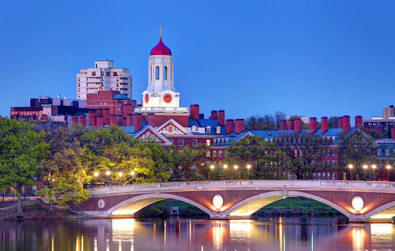 Dusk skyline of Cambridge, Massachusetts, the red cupola of Harvard's Dunster House rising over brick riverfront buildings with the lit Weeks Footbridge spanning the Charles River in the foreground